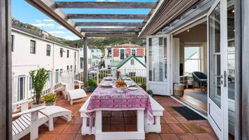 A covered outdoor dining area with a table set for two, white chairs, and potted plants.