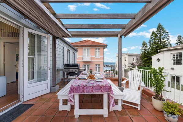 An outdoor patio with a table set for dining, a grill, and a view of a pink building and trees.