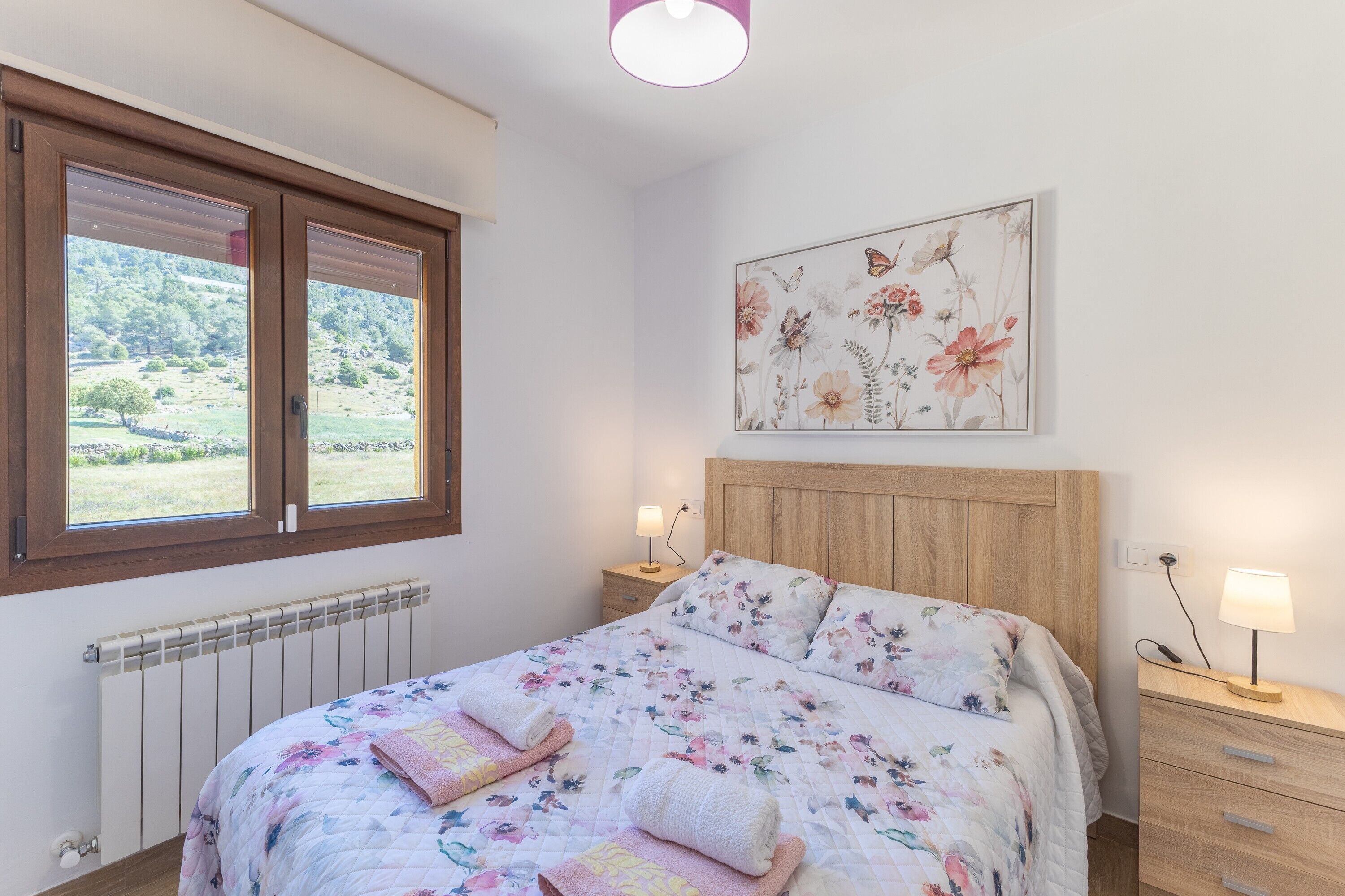 Een slaapkamer met een houten hoofdbord, een bloemenbedovertrek, twee nachtkastjes met lampen en een raam met zonwering.