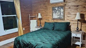 Un dormitorio con piso de madera, una cama con colcha estampada, dos mesitas de noche con lámparas, una ventana con cortinas y un equipo de aire acondicionado en la pared.