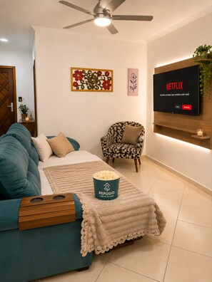 Uma sala de estar com um sofá verde-água, uma poltrona estampada, uma mesa de centro com um balde de pipoca, um ventilador de teto, uma TV fixada na parede e uma obra de arte floral emoldurada.