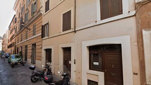 Rua estreita com scooters estacionadas e um carro verde. Edifícios com várias janelas e portas.