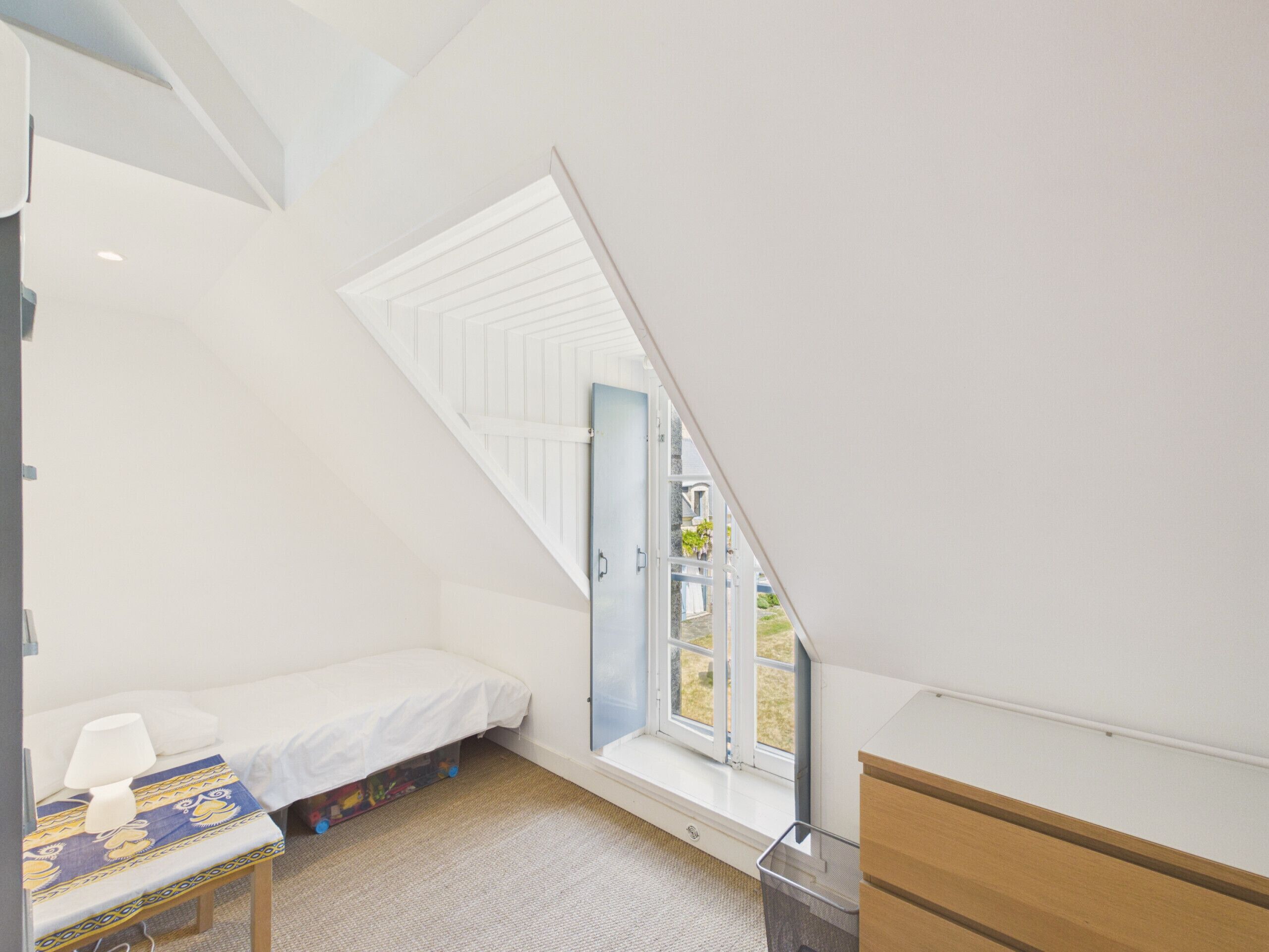 Ein kleines, dreieckiges Schlafzimmer im Dachgeschoss mit einem Bett, einem Fenster und einer hölzernen Kommode.