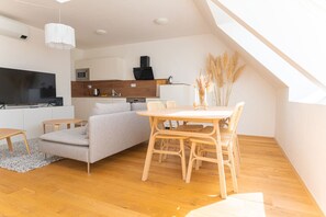 Ein modernes Wohnzimmer mit einer Couch, einem Esstisch mit Stühlen, einem Flachbildfernseher und einer Küche im Hintergrund.