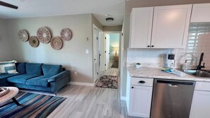 A living room with a blue sofa, a patterned rug, and wall decor. A kitchen area with white cabinets and a dishwasher is visible. - Chill Vibes B - 2 min to Beach (Fort Myers Beach)