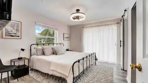 A bedroom with a bed, bedside table, lamp, chair, and a view of the outdoors through the window. - West Palm Beach 3BR Luxury Home Near Beach&Dining (West Palm Beach)