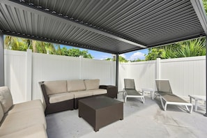 An outdoor patio with a covered seating area, including a sofa and chairs, and a white fence in the background. - West Palm Beach 3BR Luxury Home Near Beach&Dining (West Palm Beach)
