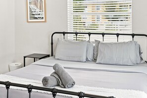 Un lit avec un cadre en métal, une literie blanche et deux oreillers. Il y a une petite table de chevet à côté, sur laquelle se trouve un rouleau de papier toilette.