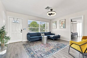 Un salon moderne avec un canapé bleu, un fauteuil jaune, un tapis à motifs et un ventilateur de plafond.