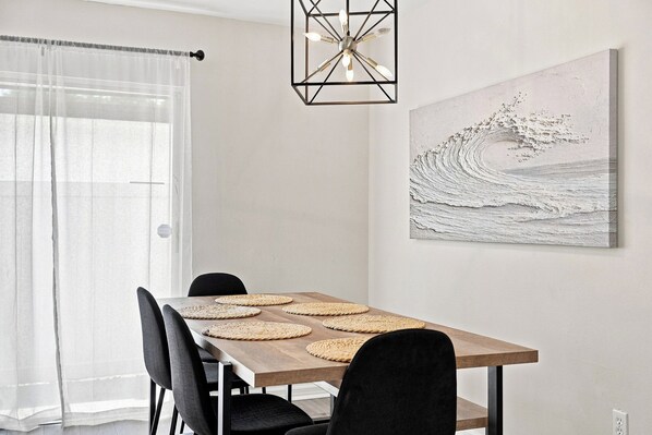 Un coin repas avec une table en bois, des chaises noires, des sets de table en fibres tressées, un lustre moderne et une œuvre d’art murale à la texture intéressante.