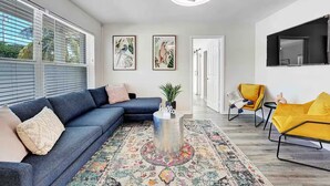 A modern living room with a sectional sofa, a yellow armchair, a coffee table, and a large window with blinds. - West Palm Beach 3BR Luxury Home Near Beach&Dining (West Palm Beach)
