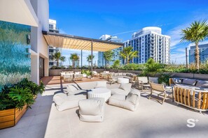 A rooftop patio with outdoor seating, palm trees, and modern buildings in the background. - Brand-New Luxury 1BR in Crosby - Rooftop pool (Miami)