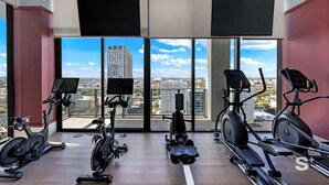 Ein modernes Fitnessstudio mit verschiedenen Kardio-Geräten, großen Fenstern und Stadtblick sowie an Wänden angebrachten Fernsehern.