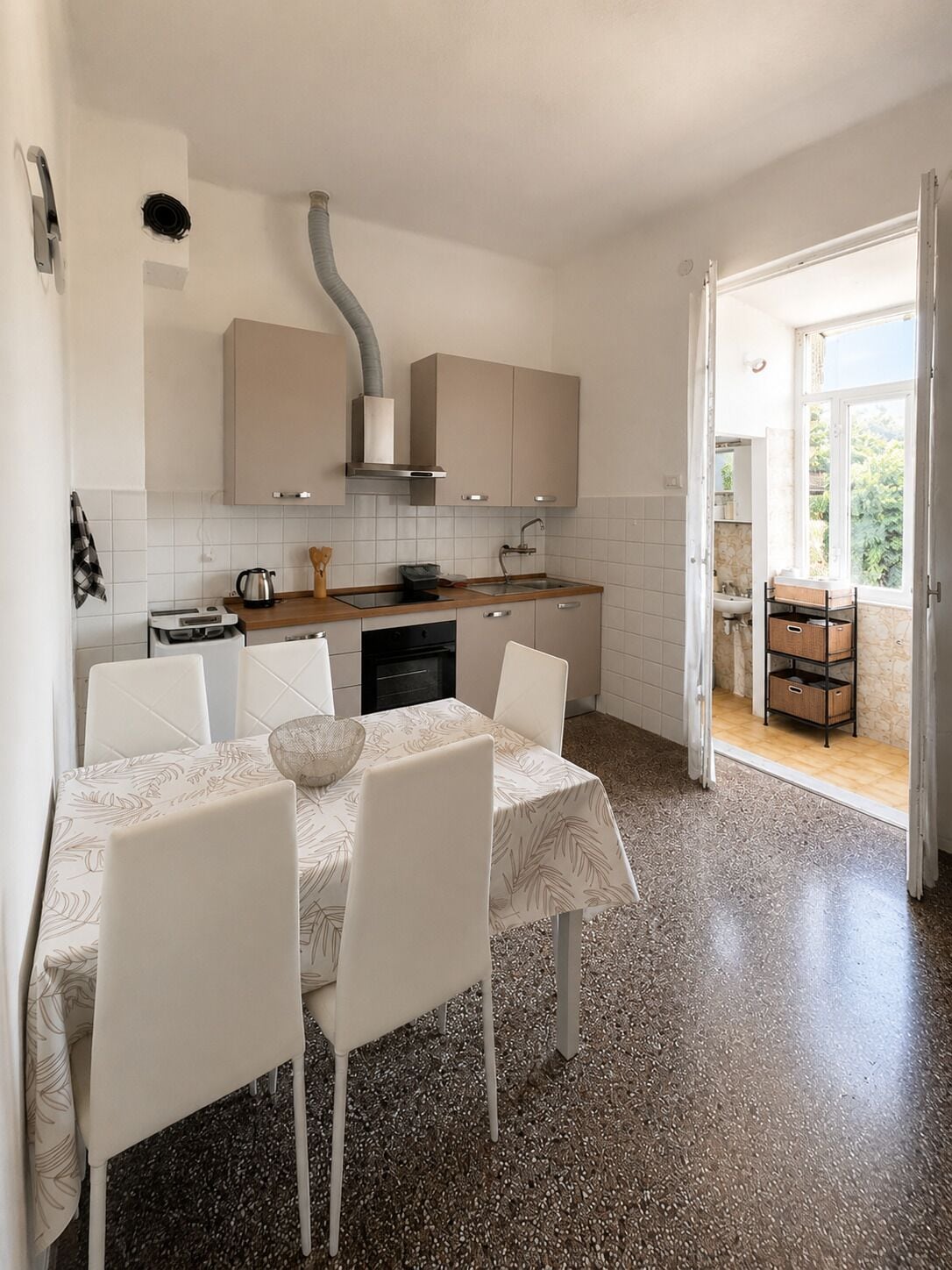 Une cuisine moderne avec des meubles blancs, une table à manger avec des chaises et, vue par une porte ouverte, un balcon.