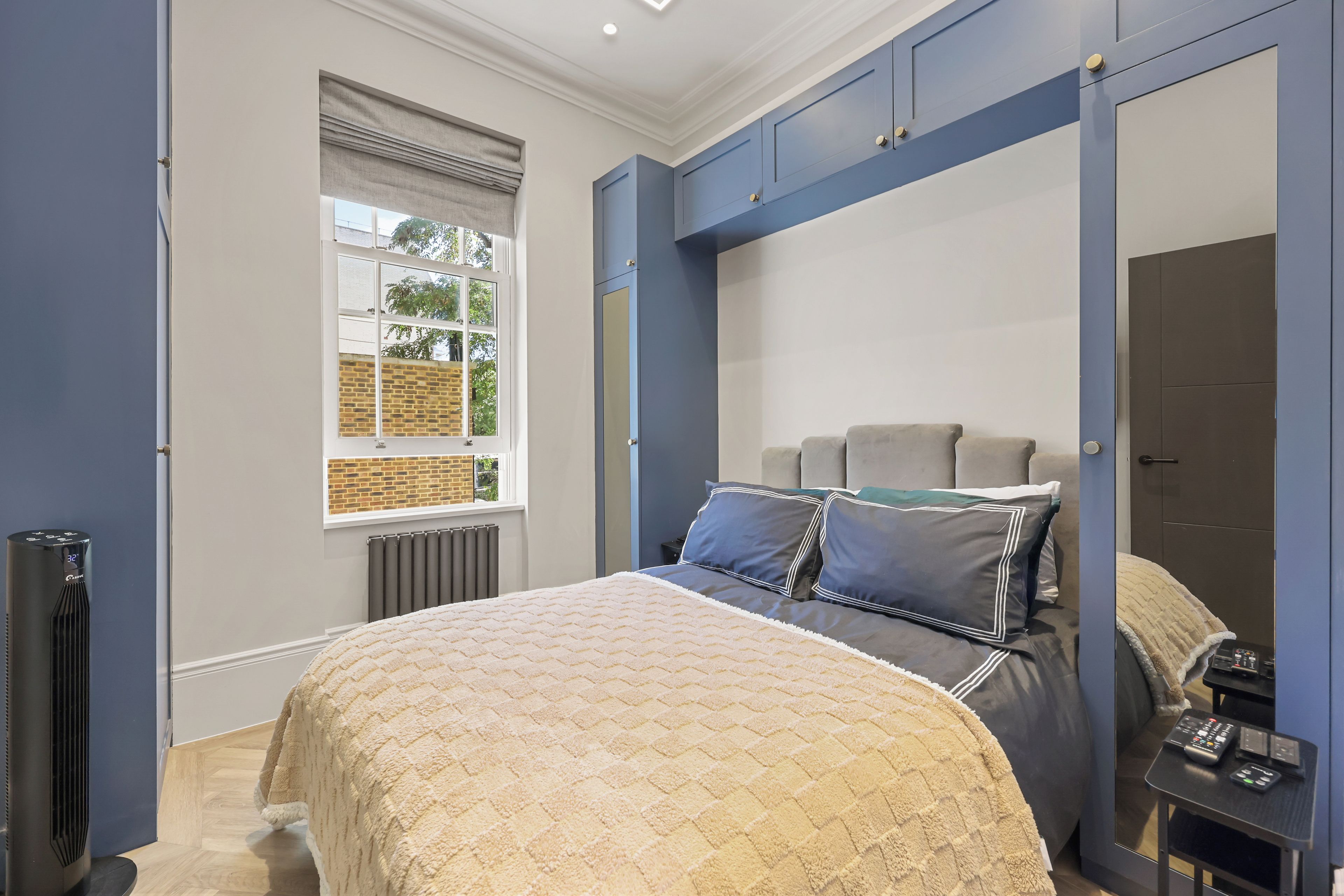 A bedroom with a bed, a window, a radiator, a heater, and a bedside table with a remote control.