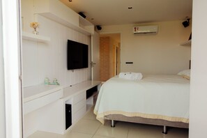 Une chambre à coucher avec un lit, un téléviseur fixé au mur blanc, une petite étagère avec des objets décoratifs et un appareil de climatisation.
