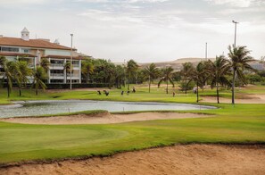 Um campo de golfe com bunkers, um lago, palmeiras e um edifício com uma torre.