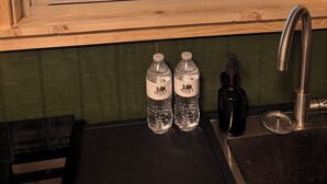 A window with a view of a wooden deck and trees, a sink with a faucet, and two water bottles labeled 'Duck's Cabin.' - Waterfront Cabin in Hilliard, FL (Hilliard)