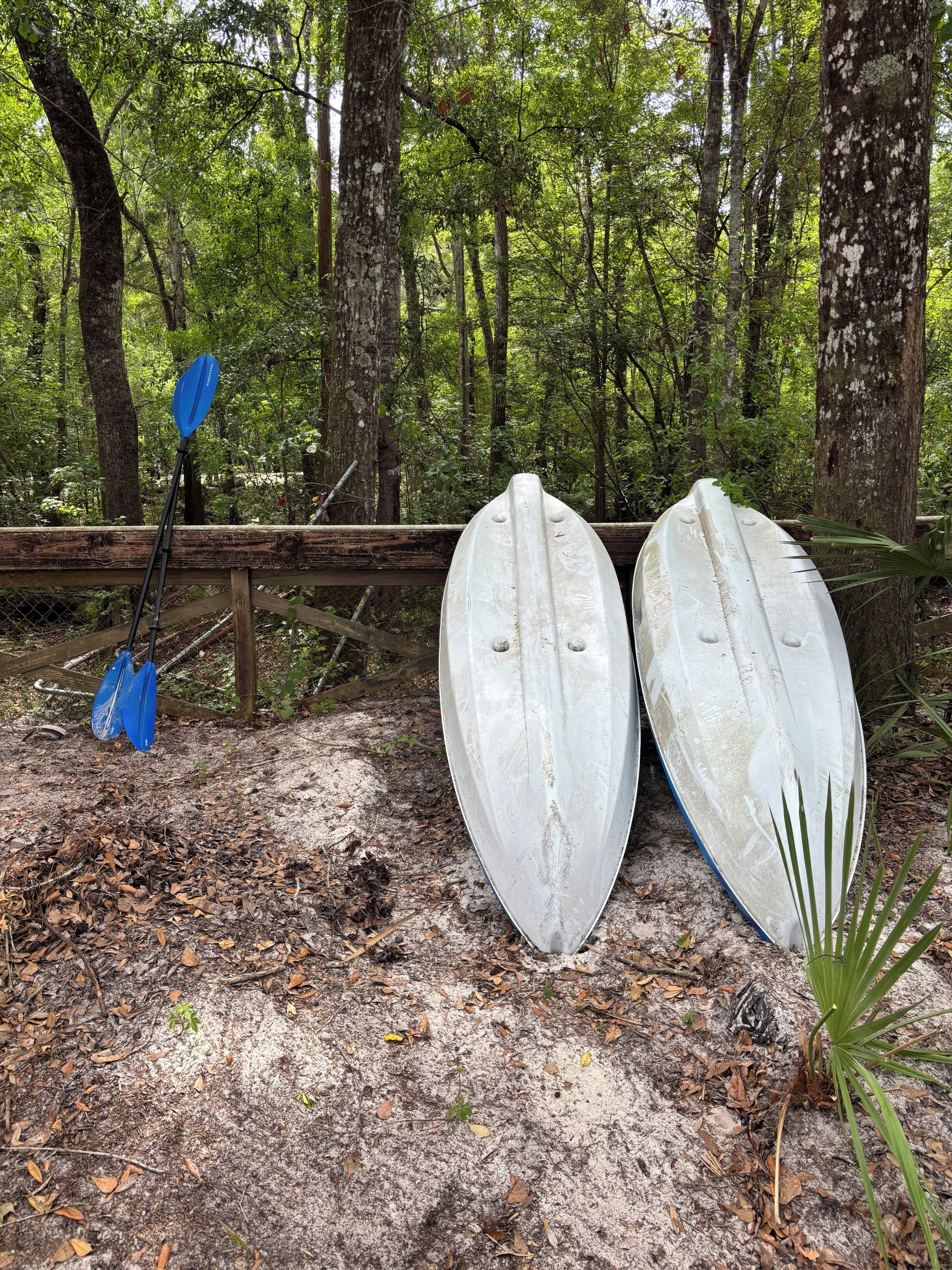 Deux kayaks blancs et une pagaie bleue sont appuyés contre une clôture en bois, dans une zone boisée.