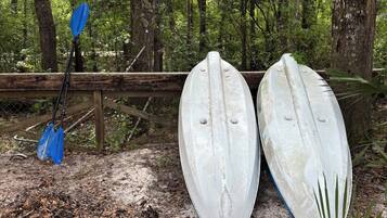 Deux kayaks blancs et une pagaie bleue sont appuyés contre une clôture en bois, dans une zone boisée.