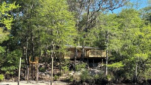A wooden cabin with a deck, surrounded by dense trees and a body of water. - Waterfront Cabin in Hilliard, FL (Hilliard)