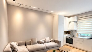 A modern living room with a sectional sofa, a round coffee table, and a kitchen area with a stove and cabinets. - Luxury Apartament (Tiranë)