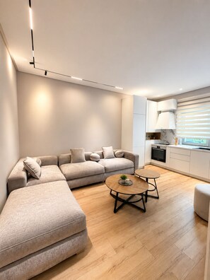 A modern living room with a sectional sofa, a round coffee table, and a kitchen area with a stove and cabinets. - Luxury Apartament (Tiranë)
