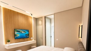 A modern bedroom with a large bed, a TV mounted on a wooden panel, and a glass door leading to a bathroom. - Luxury Apartament (Tiranë)