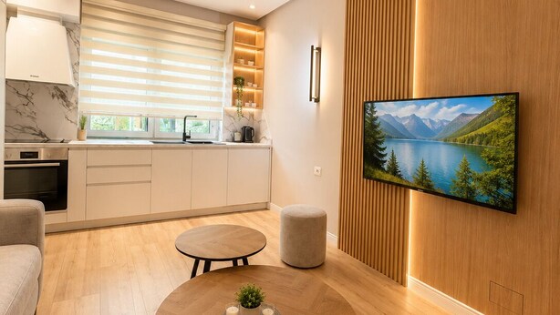 A modern living room with a round coffee table, a sofa, a TV mounted on a wooden wall, and a kitchen in the background. - Luxury Apartament (Tiranë)