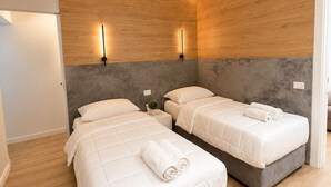 A modern hotel room with two beds, a grey headboard, wooden panel walls, and a small bedside table with a plant. - Luxury Apartament (Tiranë)