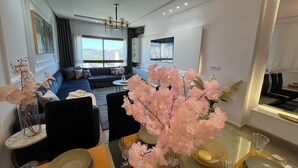 A modern living room with a sofa, a dining table set for two, and a large window offering a city view. - 2-bedroom apartment in adorable Tangier with AC comfort (Tangier)