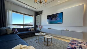 A modern living room with a sofa, two small round tables, a large TV on the wall, and a view of the city. - 2-bedroom apartment in adorable Tangier with AC comfort (Tangier)