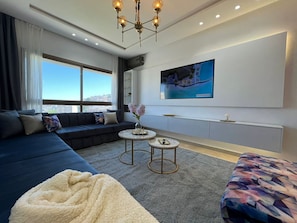 Ein modernes Wohnzimmer mit einer Couch, zwei kleinen runden Tischen, einem großen Fernseher an der Wand und Blick auf die Stadt.
