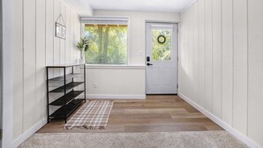 Un couloir avec un plancher en bois, une porte blanche ornée d’une guirlande, une petite étagère et une fenêtre donnant sur des arbres.
