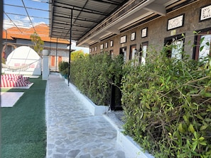 A covered outdoor area with a stone pathway, green artificial turf, and a row of windows with decorative frames. - Nusa Penida Homestay (Penida Island)