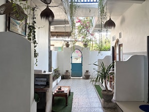 A narrow indoor corridor with hanging plants, a laptop on a table, and a blue door at the end. - Nusa Penida Homestay (Penida Island)
