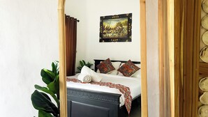 A bedroom with a large mirror, a bed with patterned bedding, a painting on the wall, and a plant. - Nusa Penida Homestay (Penida Island)