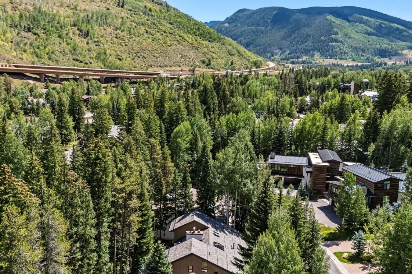 A residential area with a modern house surrounded by dense forest, a bridge in the background, and a mountain range.
