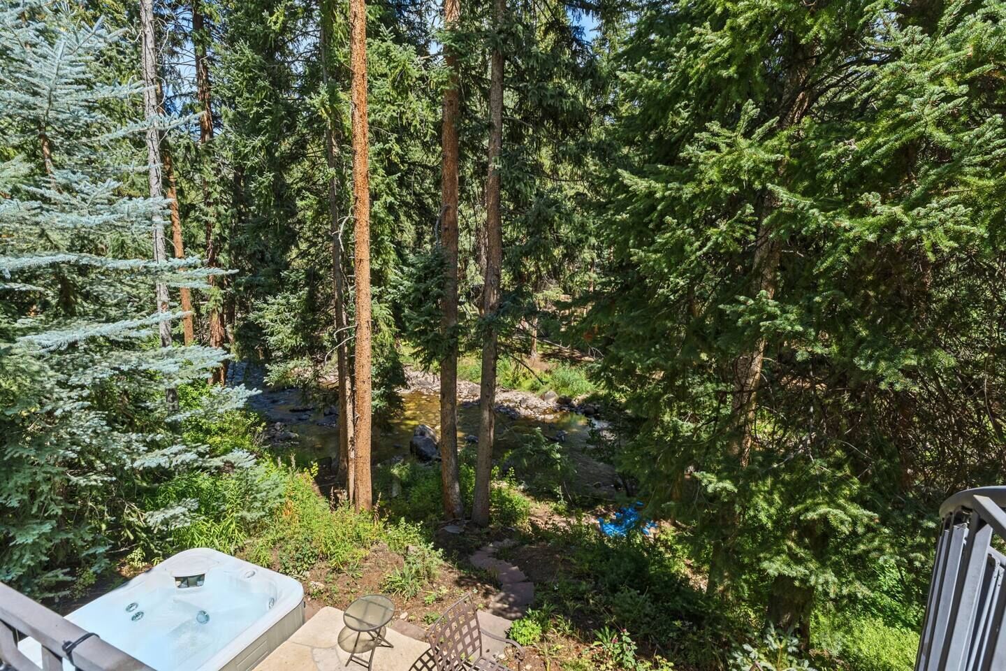 A patio area with a hot tub, outdoor seating, and a view of a forested area with a stream.