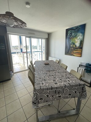 Un comedor con mantel estampado, sillas, ventilador de techo, un cuadro y vista a través de un vidrio.