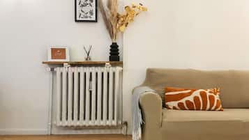 A room with a radiator, a framed picture, a vase with dried plants, and a sofa with a patterned pillow.