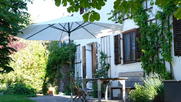Un patio con una mesa y sillas, una sombrilla blanca y un edificio de color blanco con contraventanas de madera.