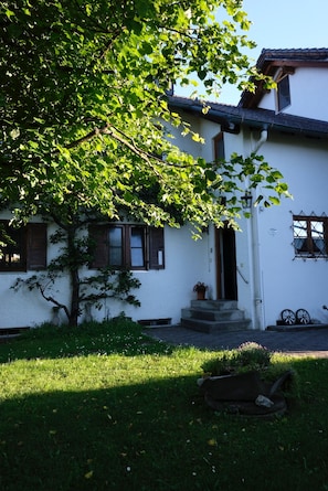 Un edificio blanco con un árbol en frente, un pequeño porche y una ventana con contraventanas.