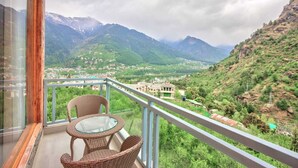 Ein Balkon mit Korbmöbeln und einem Glastisch mit Blick auf eine gebirgige Landschaft und Gebäude.
