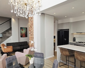 Eine moderne Küche mit einem schwarzen Kühlschrank, ein Essbereich mit einem Glastisch und Stühlen, ein Wohnzimmer mit einer Couch und einer Chandelier.