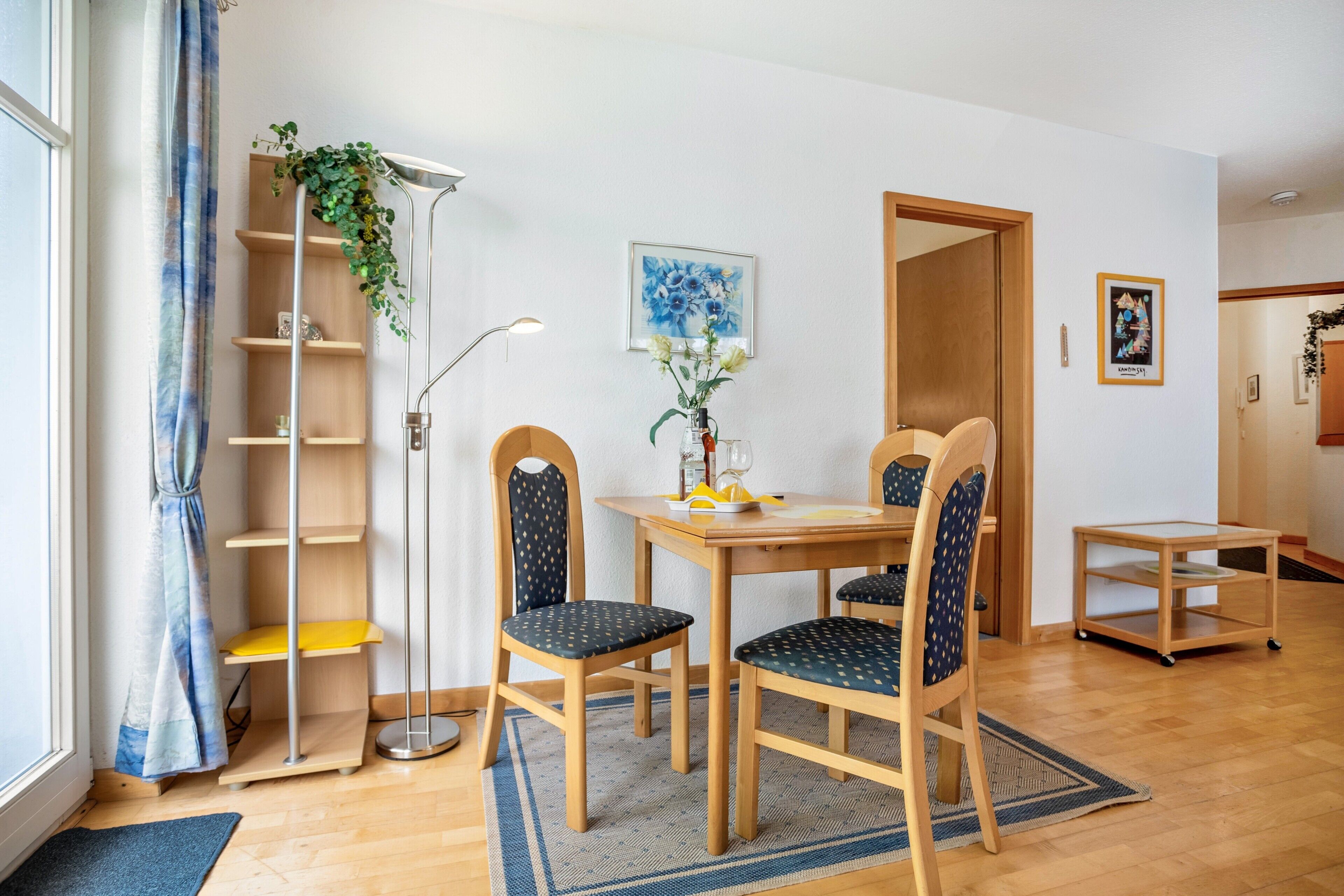 A room with a wooden dining table, two chairs, a floor lamp, a bookshelf, and a small table with a plant.