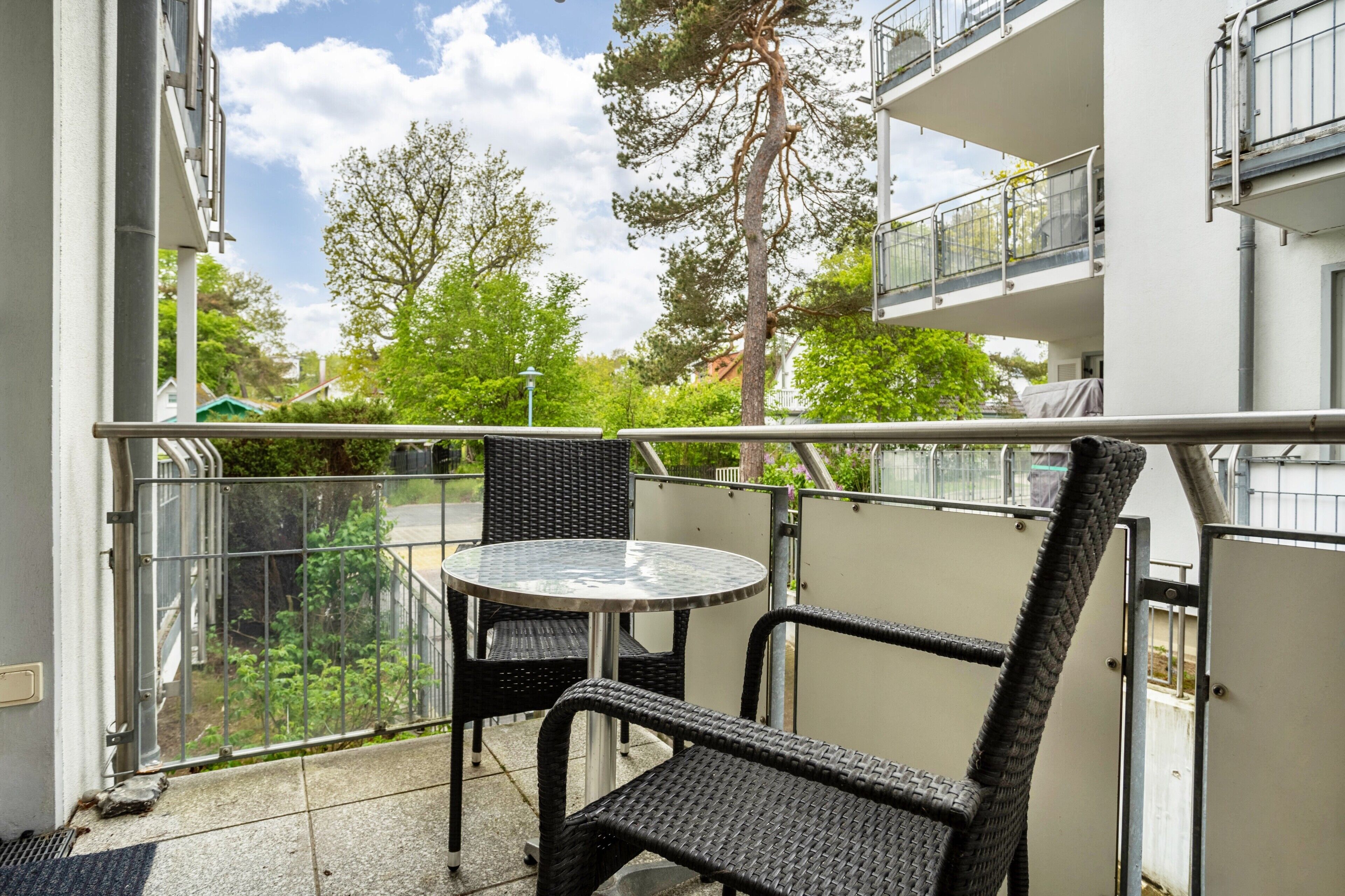 Een balkon met een ronde tafel en twee stoelen, met uitzicht op een woongebied met bomen en gebouwen.