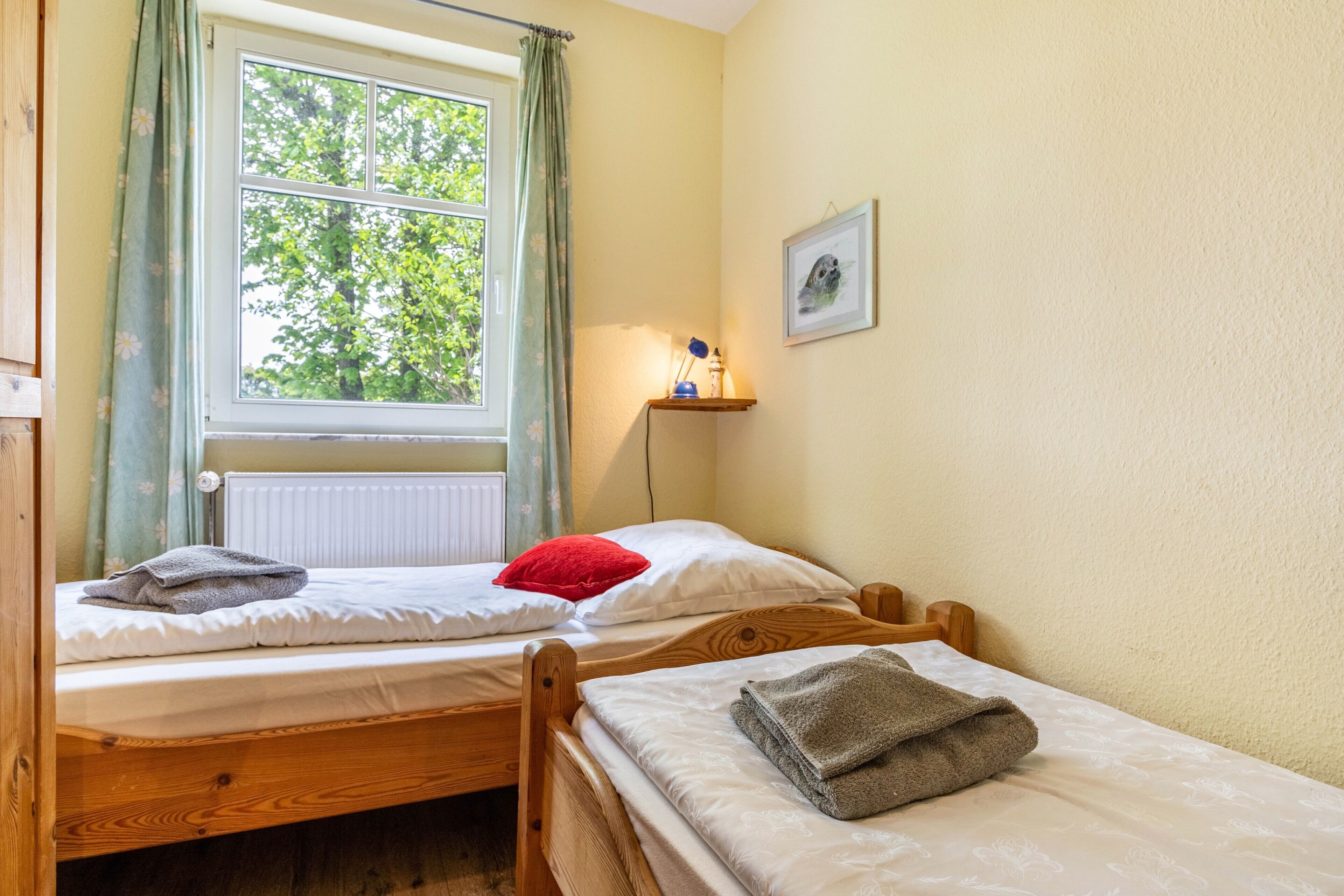 Een kamer met twee houten bedden, een raam met gordijnen, een radiator, een klein plankje met een lamp en een ingelijst beeld aan de muur.
