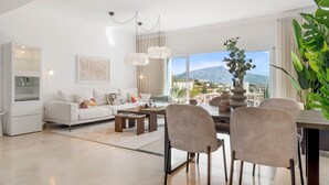 En moderne stue med sofa, sofabord og spiseplads, store vinduer med udsigt til et bjerglandskab, og en plante.