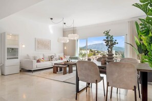 A modern living room with a sofa, coffee table, and dining area, large windows with a mountain view, and a plant. - Luxurious Penthouse - View, Roof Terrace, 3BR/2BA (Benahavís)
